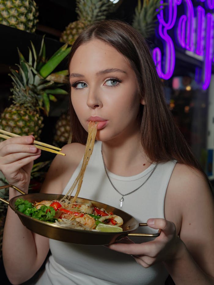 Young woman savoring an Asian noodle dish with chopsticks, surrounded by pineapples at night market.